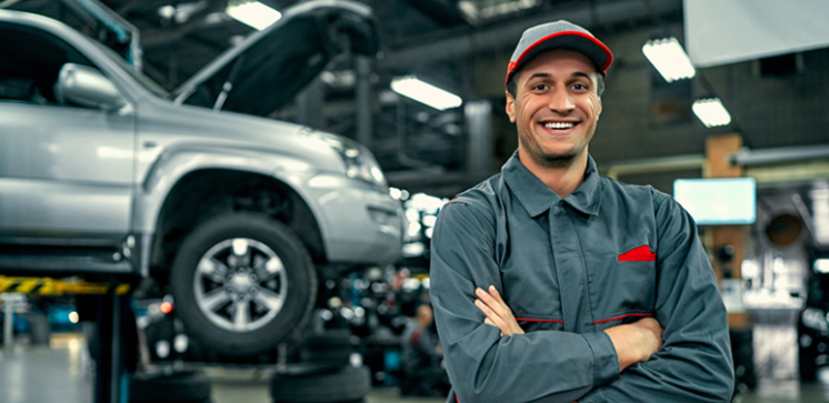 Smiling auto repair customer service advisor standing in a workshop with a vehicle lifted on a car lift in the background.