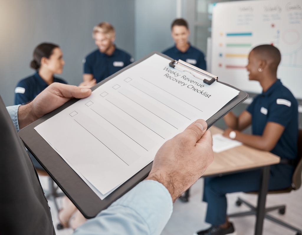 β’ n automotive service advisor holds a printed weekly revenue recovery checklist in the foreground, with an auto shop management team collaborating around a meeting room table in the blurred background.